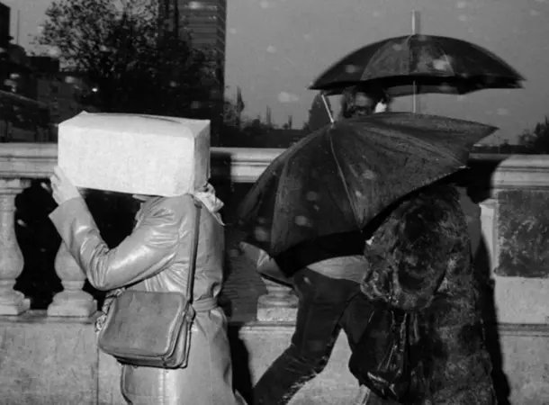 Black and white photograph showing three people walking on a bridge in rainy weather. One person covers their head with a box, while the other two use umbrellas. 