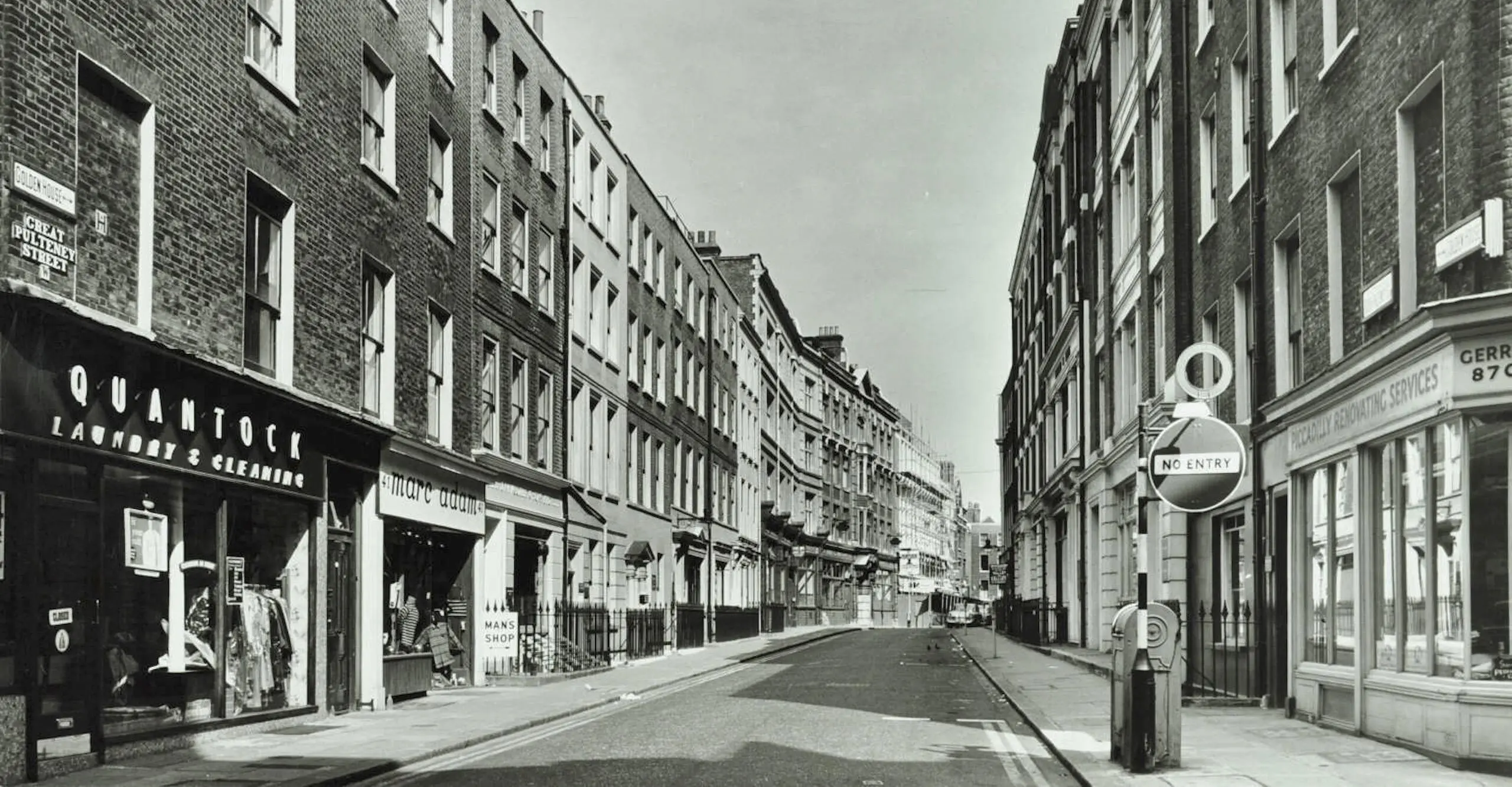 Black and white photo looking down a street with commercial shops at ground level of four story, brick buildings.